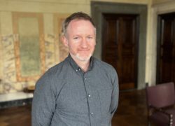 Mies, jolla on tummanharmaa kauluspaita yllään, pitää kädet selän takana ja katsoo hymyilevästi kameraan.