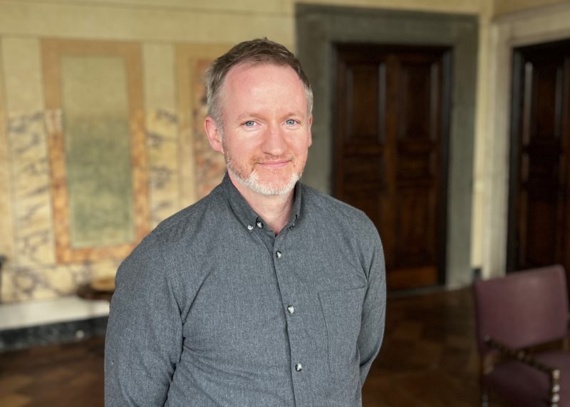 Mies, jolla on tummanharmaa kauluspaita yllään, pitää kädet selän takana ja katsoo hymyilevästi kameraan.