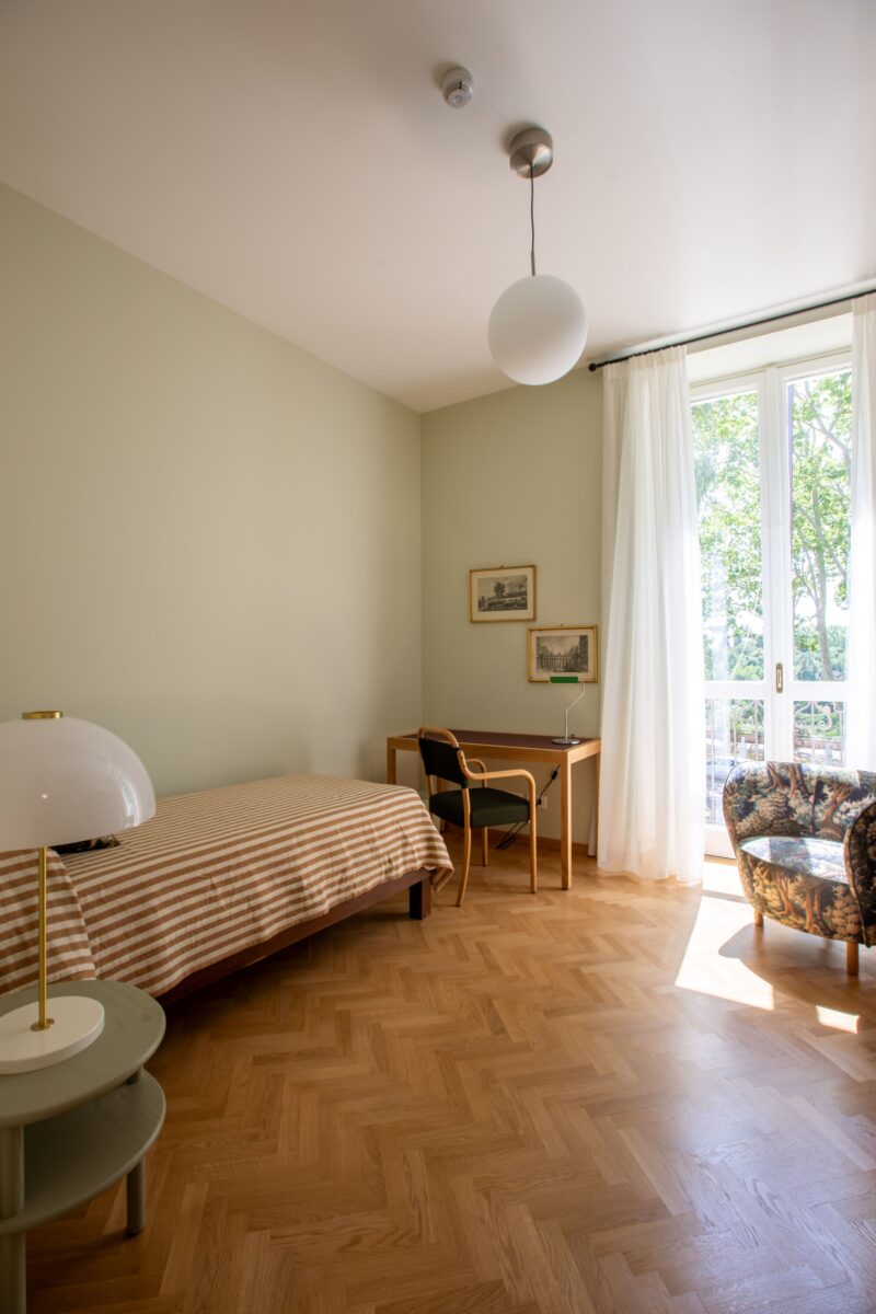 Valokuva yhden hengen residenssihuoneesta, josta löytyy sänky, työpöytä, vaatekaappi ja nojatuoli. Vaaleanvihreät seinät ja parkettilattia.