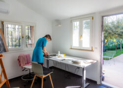 Henkilö sinisessä mekossa seisoo pöydän äärellä ja maalaa. Huoneessa maalausteline. Taustalla näkyy puutarha.