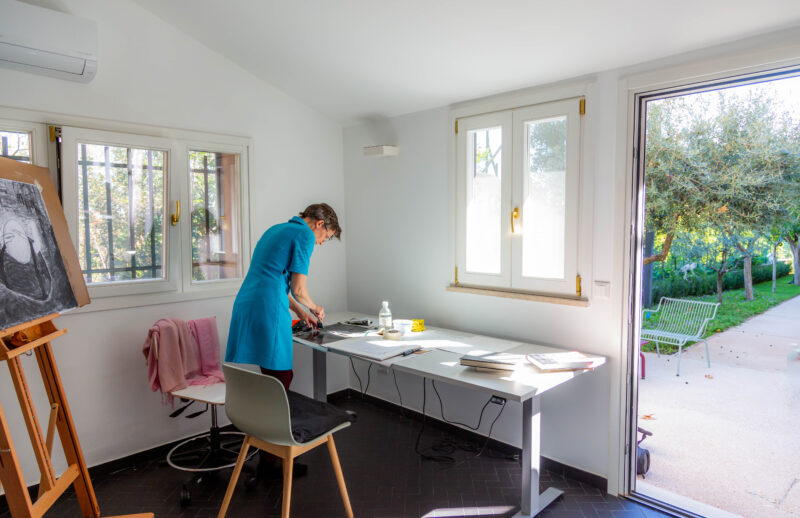 Henkilö sinisessä mekossa seisoo pöydän äärellä ja maalaa. Huoneessa maalausteline. Taustalla näkyy puutarha.